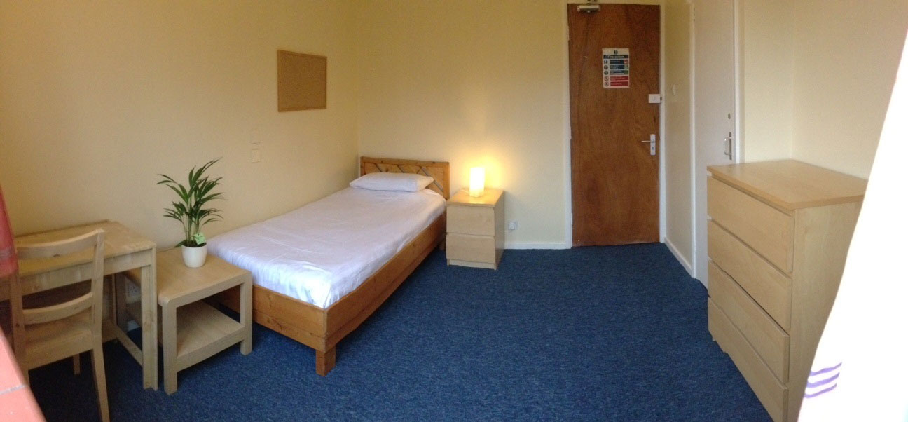 A photo of a bedroom , the bed is in the left hand corner, the door in the right, there is a chest of drawers in the front right corner of the photo. There are blue carpets and magnolia walls