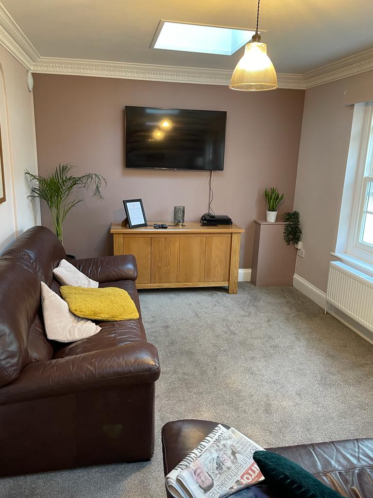 Lounge area - a room with a large brown sofa, a TV mounted on the wall, a storage unit under the tv and a window to the right of the image.