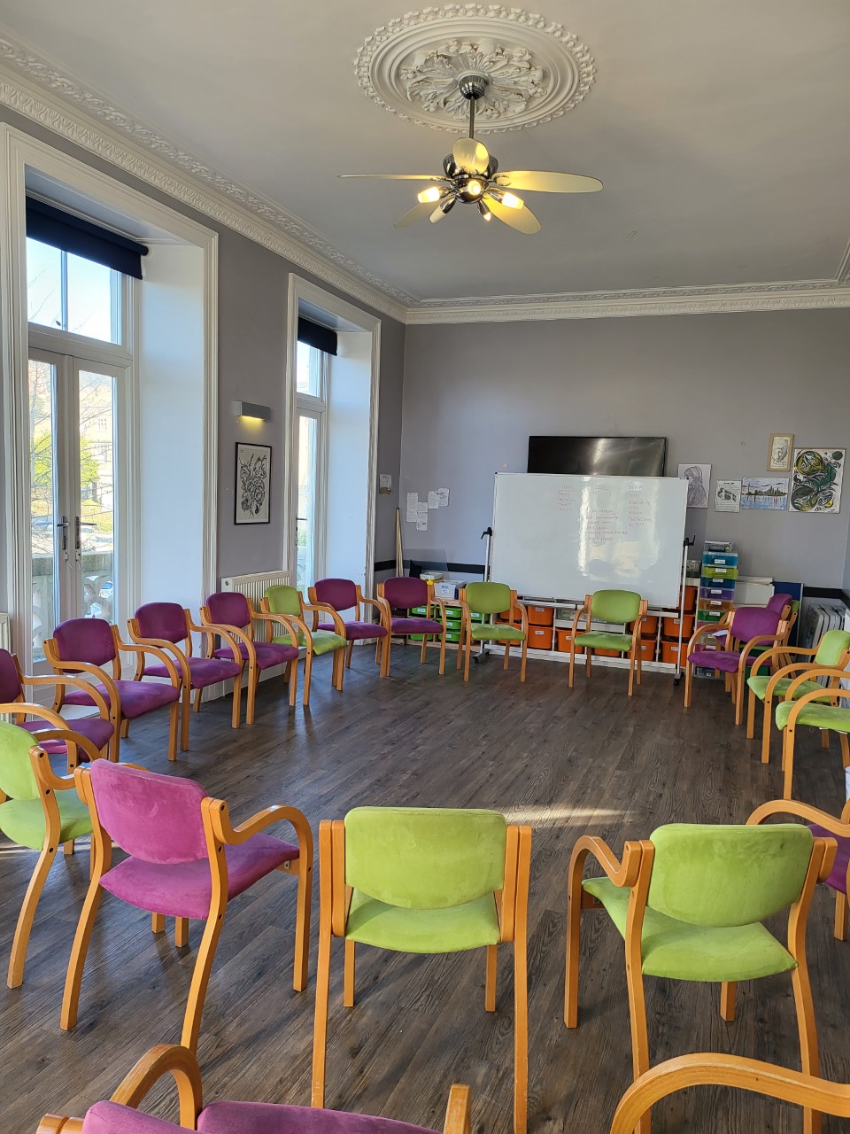 Main group room - a large room with high ceilings. a circle of padded chairs in pink and light green face the centre of the room, and there is a whiteboard on the far wall