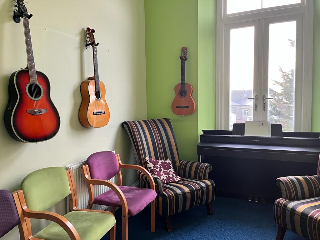 Music room - a room with three brightly coloured armchairs, and an upright piano, a large window. There are two guitars and a ukulele mounted on the wall.