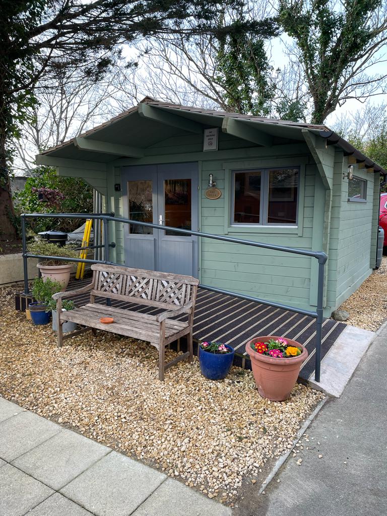 The villa - a large hut painted green. Outside the front is a gravel area with a bench and some large plant pots, and a path runs along the front