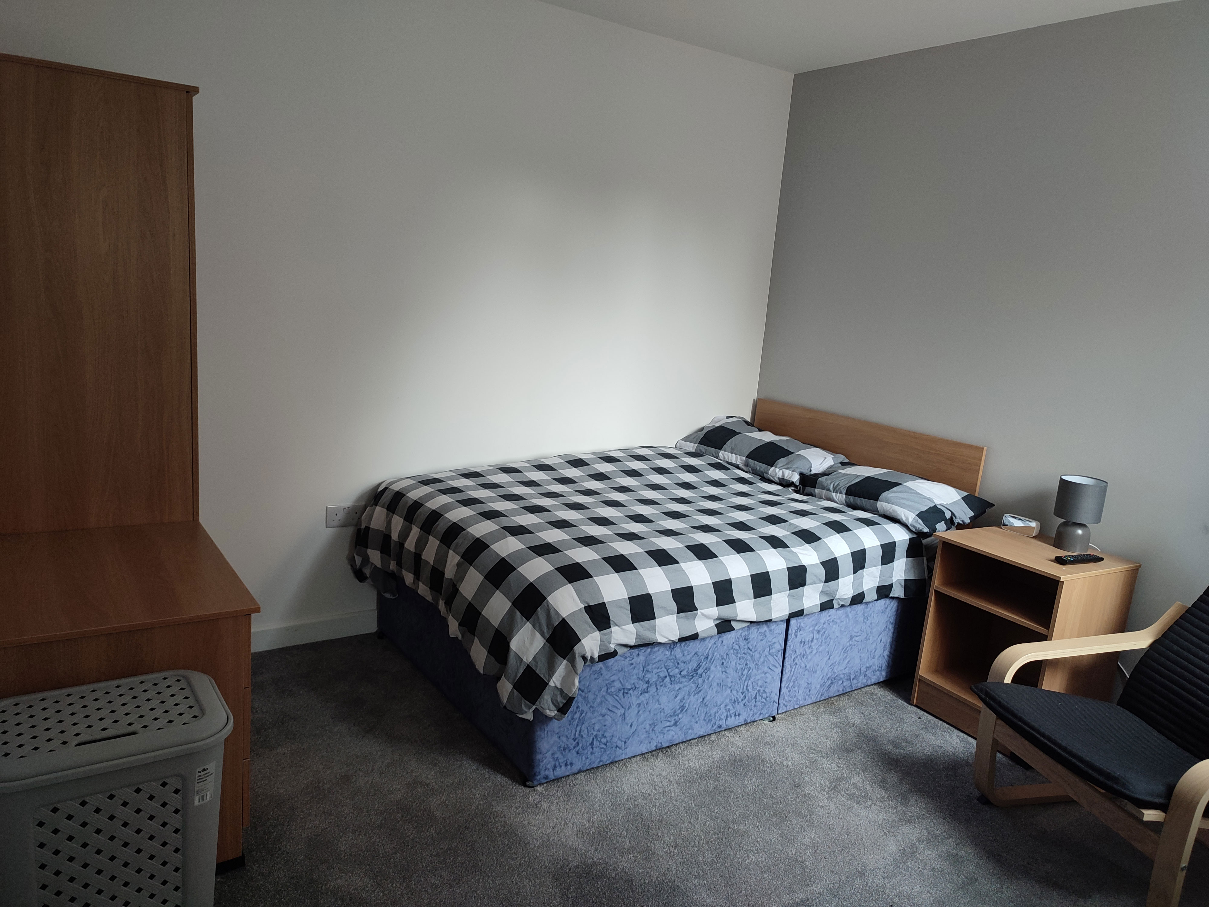 A photo of a bedroom. The room has grey walls and a grey carpet. There is a double bed in the corner of the room with a black and white gingham pattern on it. A bedside table sits next to the bed along with an arm chair. A pine wardrobe sits opposite the bed.