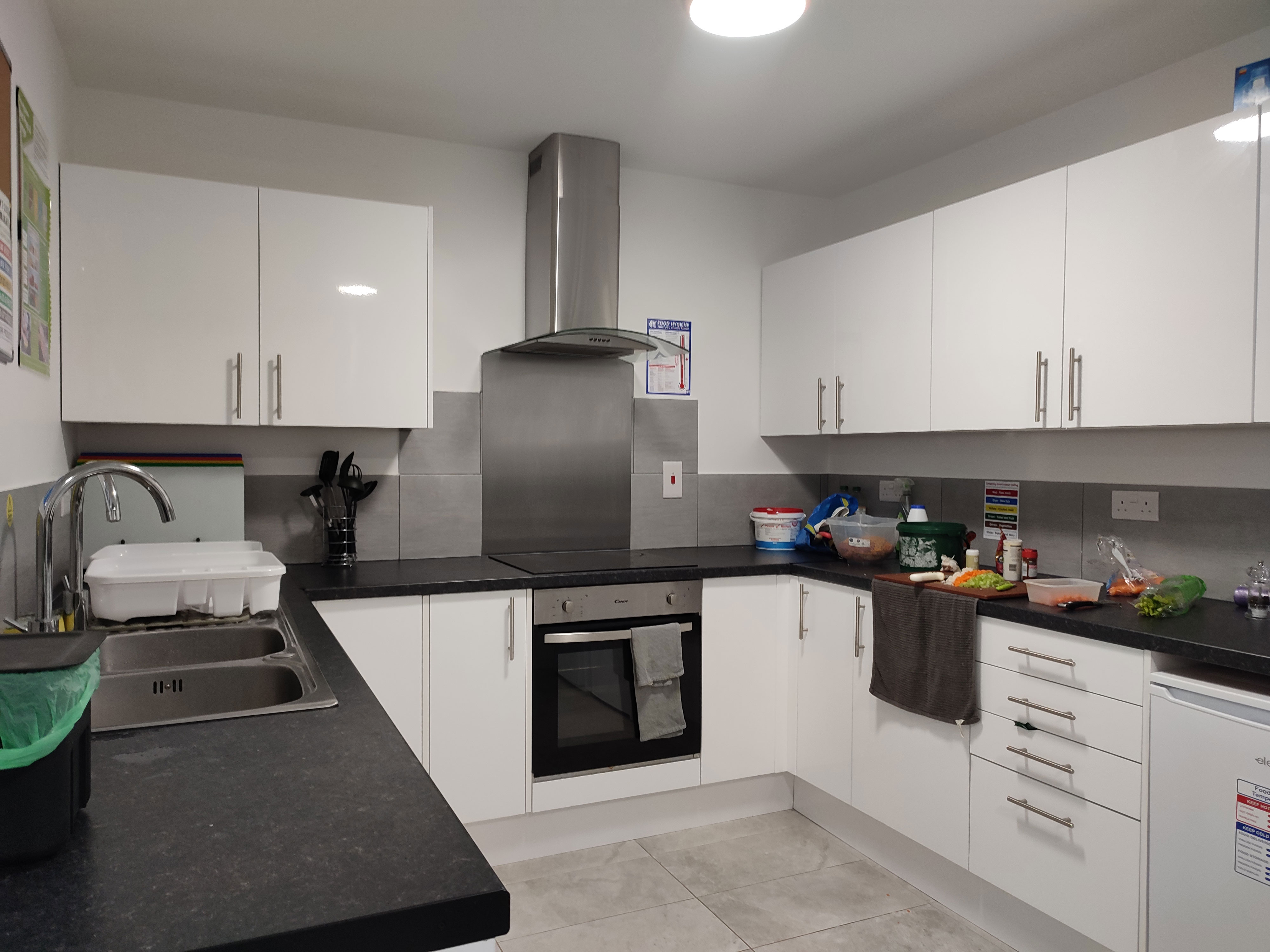 A photo of a kitchen. The cabinets and drawers are all white with silver handles. They have black countertops and a double sink. The over and stove top is on the back wall. There is a salad being prepared on the right hand side.
