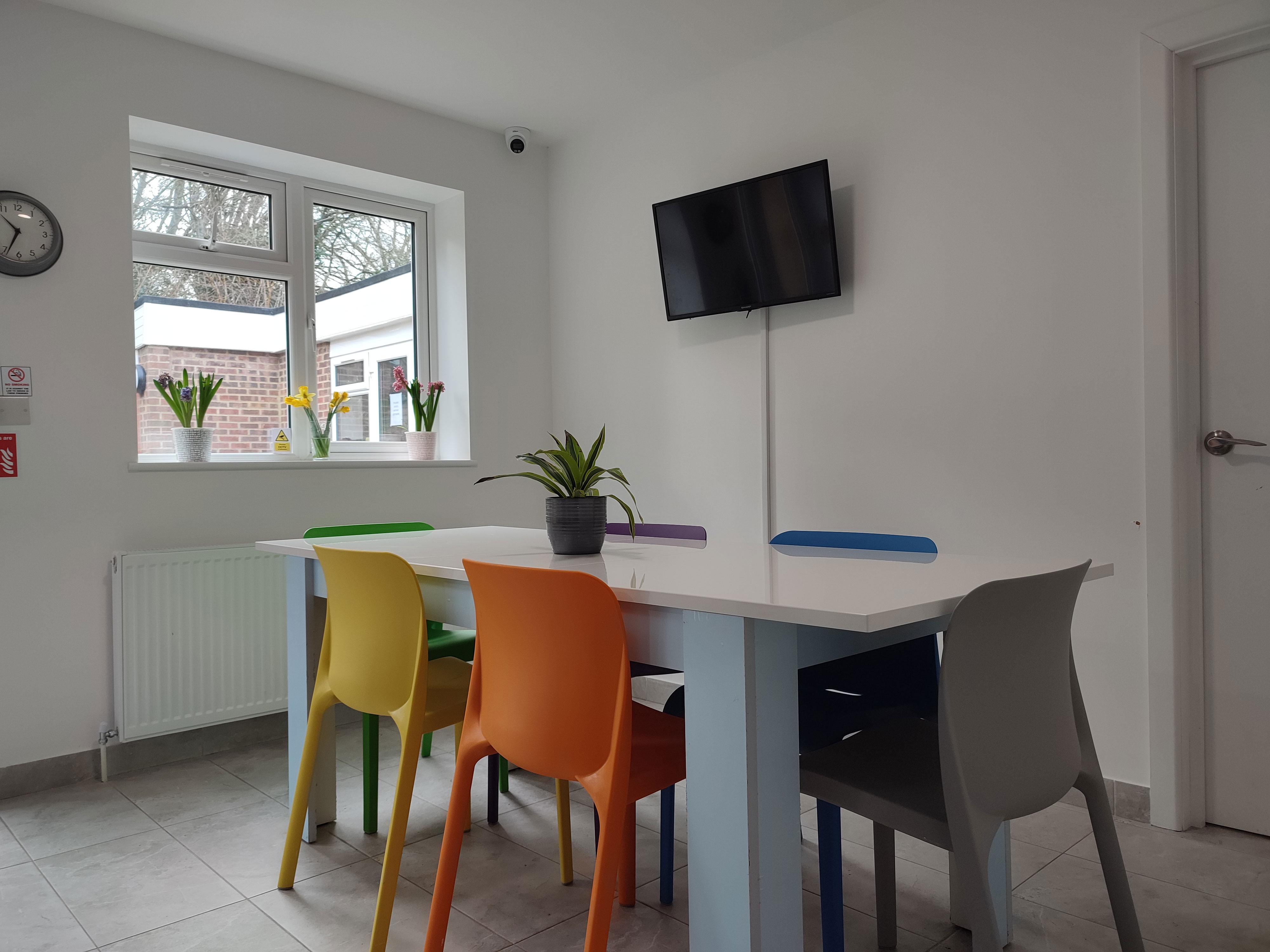 A photo of a white dining area. there is a window on the back wall and a TV on the adjacent wall. A white dining table sits in the middle with rainbow coloured plastic chairs tucked underneath.