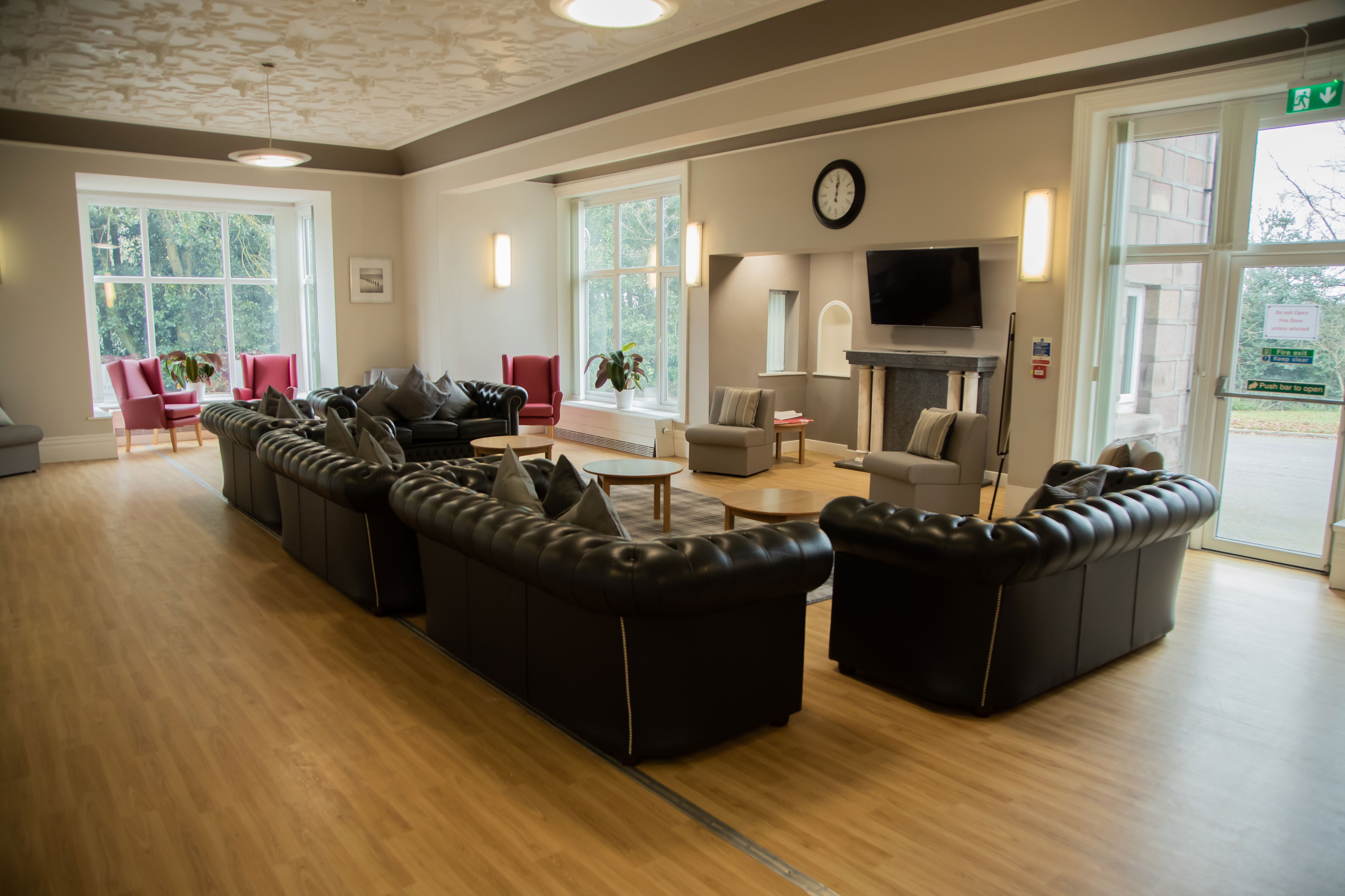 A photo of a living room. there are 5 leather sofas all facing a TV that is mounted on a wall above a fireplace There is a clock on the wall above the TV. In the corner to the room there are arm chairs in front of big windows.
