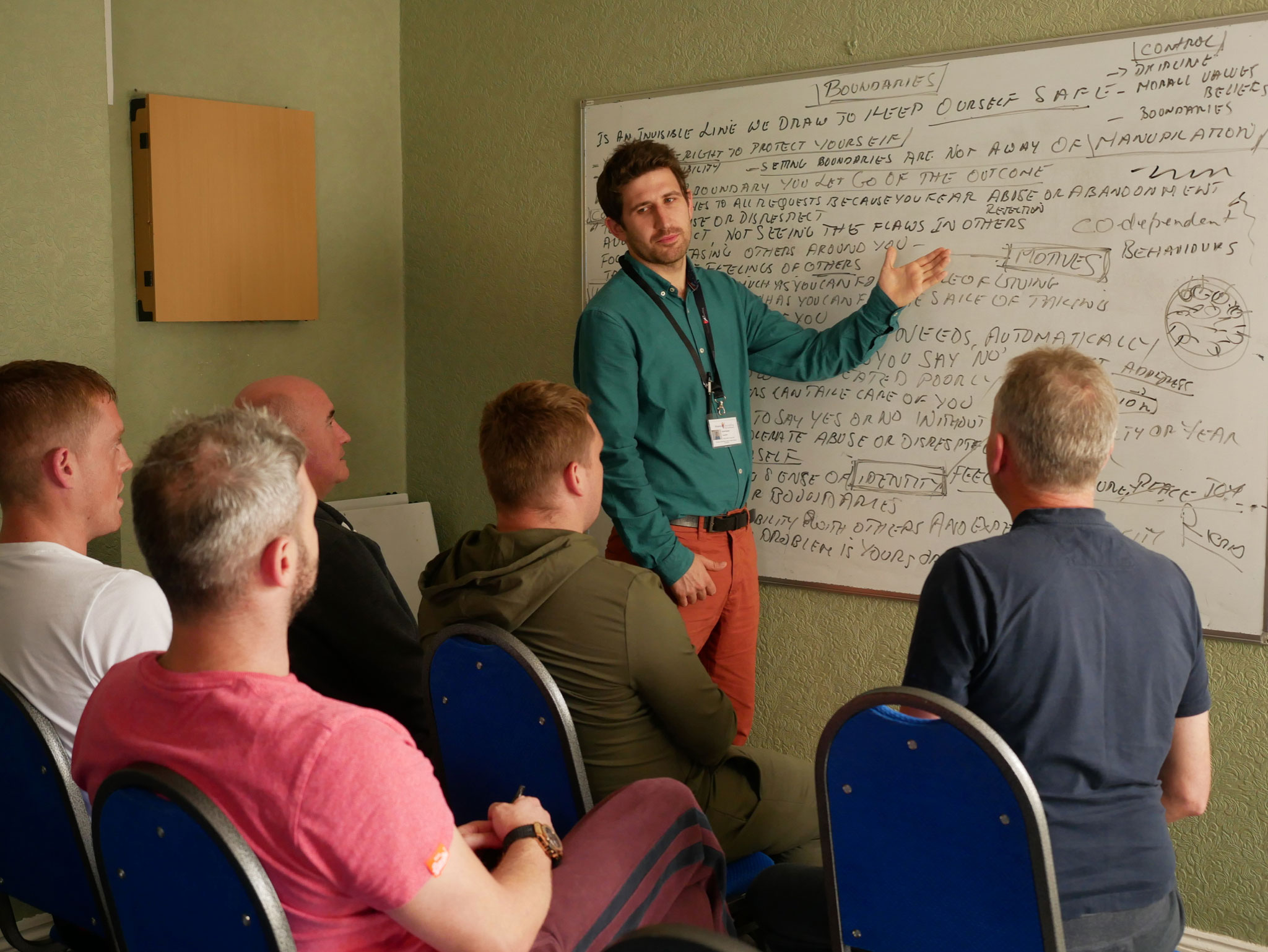 A group therapy class - with a white man standing at a whiteboard and a group of five men on chairs facing towards him