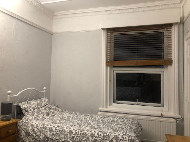 A photo of a bedroom. There is a single bed in the corner of the room with a window on the wall next to it with venetian blinds. Next to the bed is a small bedside table with a lamp on top.