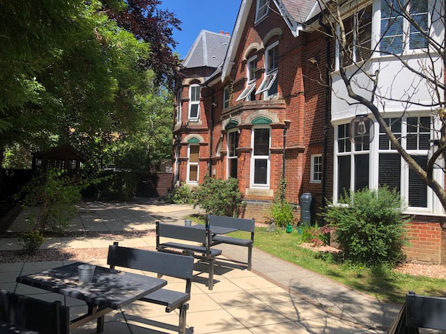A photo of a garden in front of the red brick building. There is a patio with multiple tables and chairs and plenty of trees and bushes around the edge.