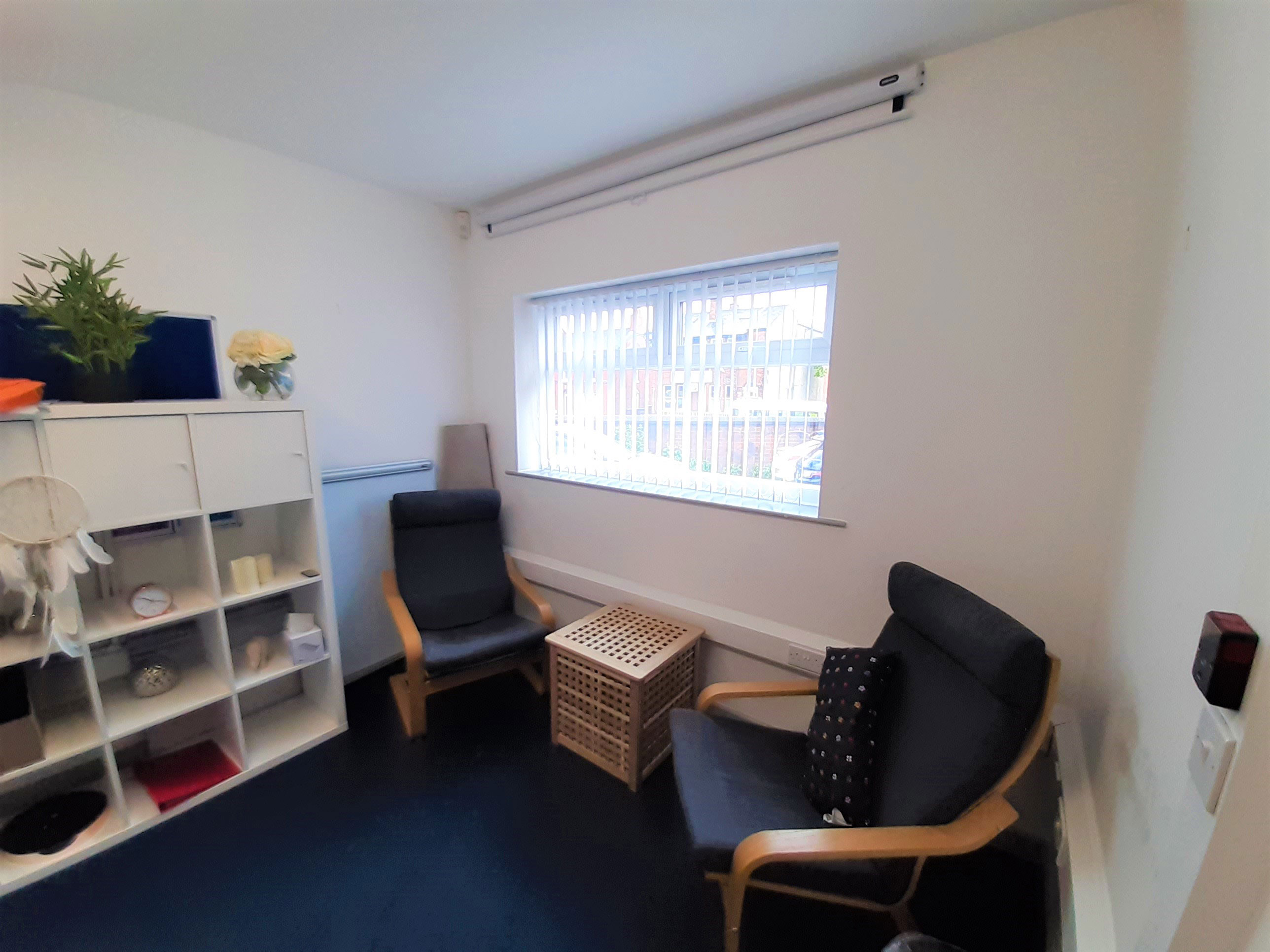 Macclesfield 121 counselling room - a room with a window, white walls, bookshelf and two chairs facing each other
