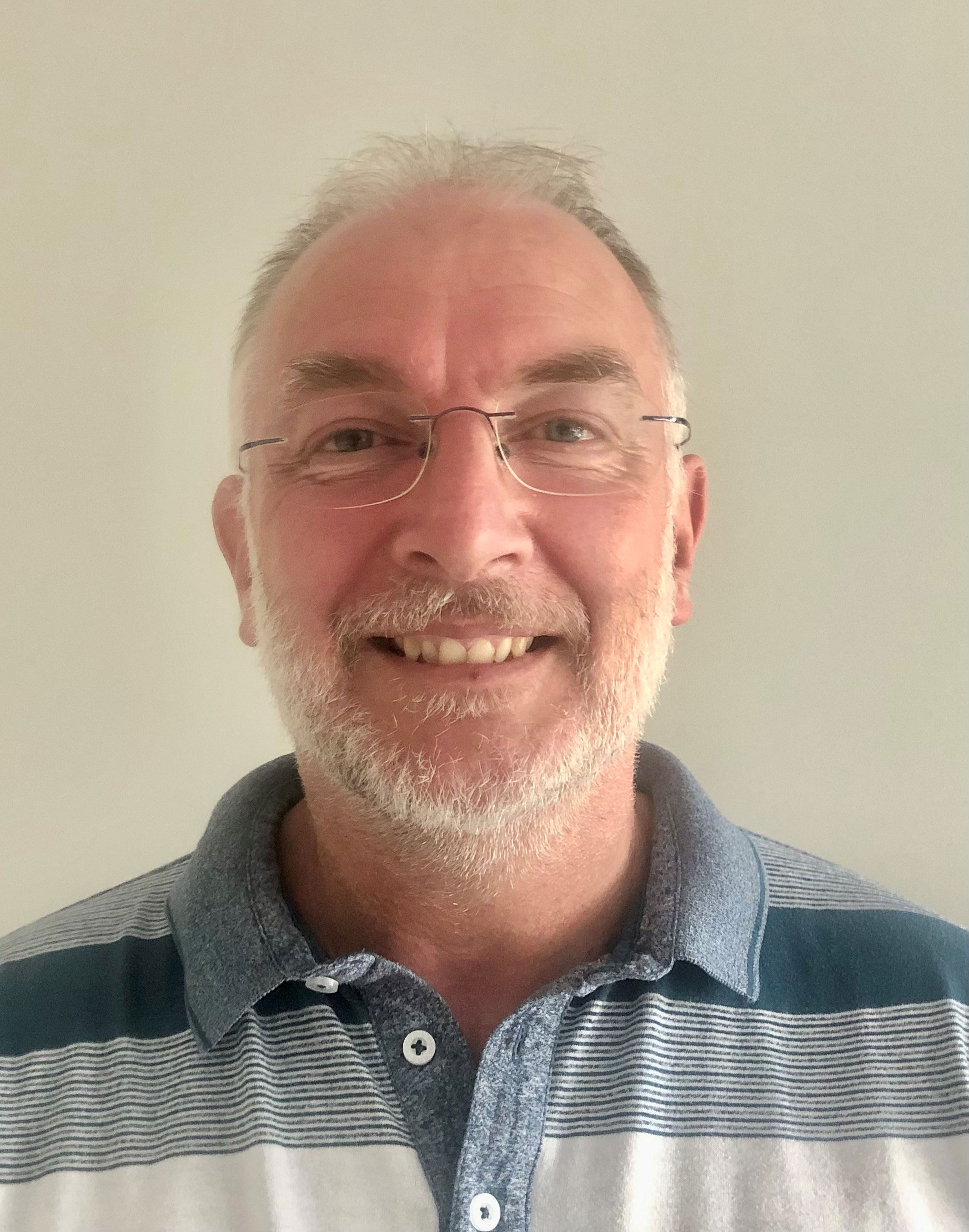 A photo of an older man smiling at the camera. He has grey hair and a grey beard. He is wearing glasses and a polo shirt with stripes of differed shades of blue and grey