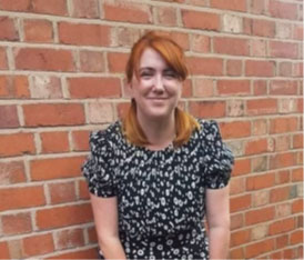 A photo of a women standing in front of a brick wall. She has ginger hair tied into two pigtails and is wearing a black top with a white pattern on