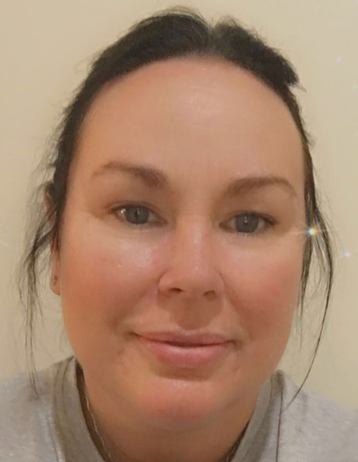 A photo of a woman smiling at the camera. She has her hair tied back from her face and is wearing a grey t-shirt