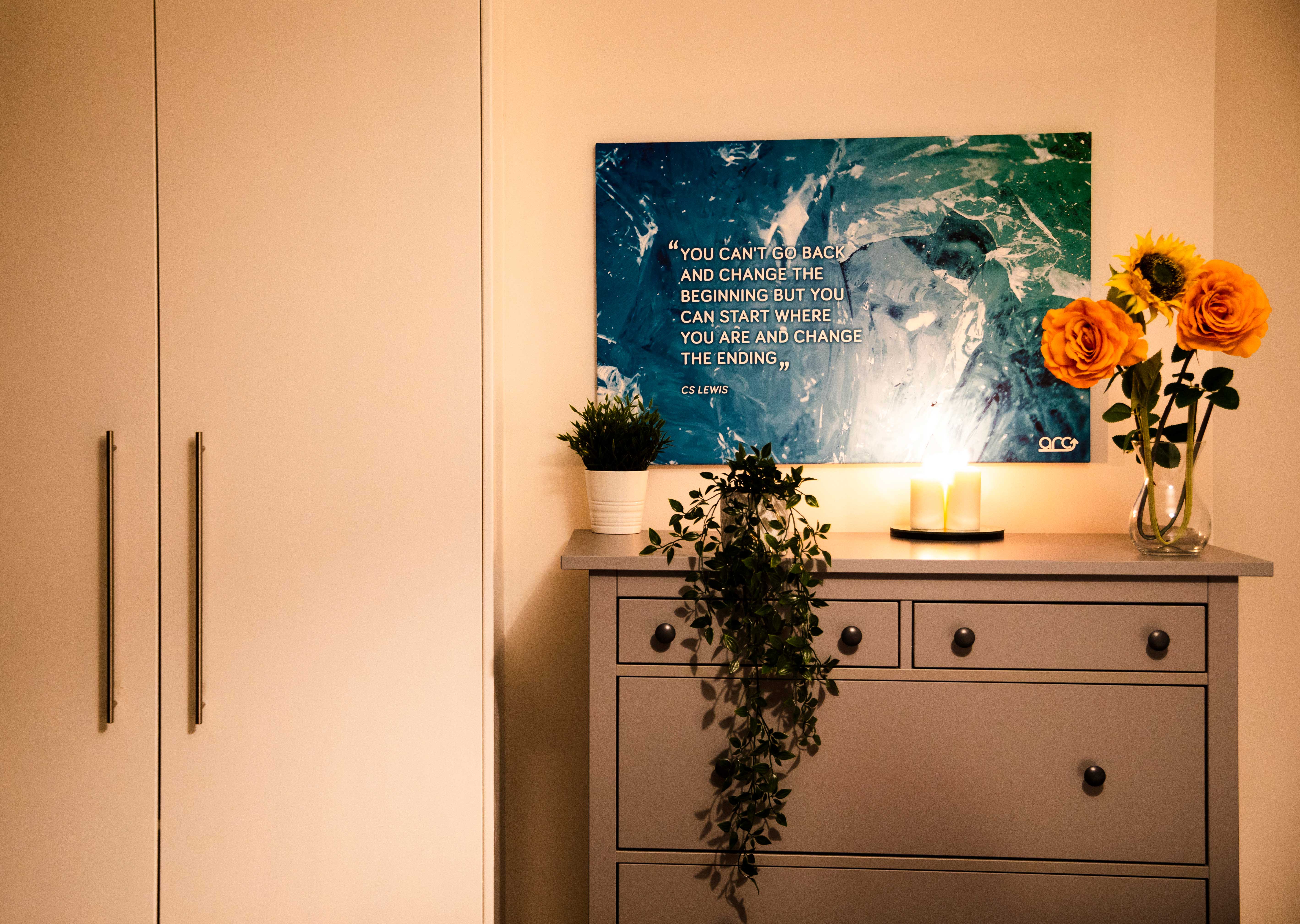a chest of drawers in the bedroom next to a wardrobe. On the chest of drawers are some fake flowers and candles. there is a piece of art on the wall.