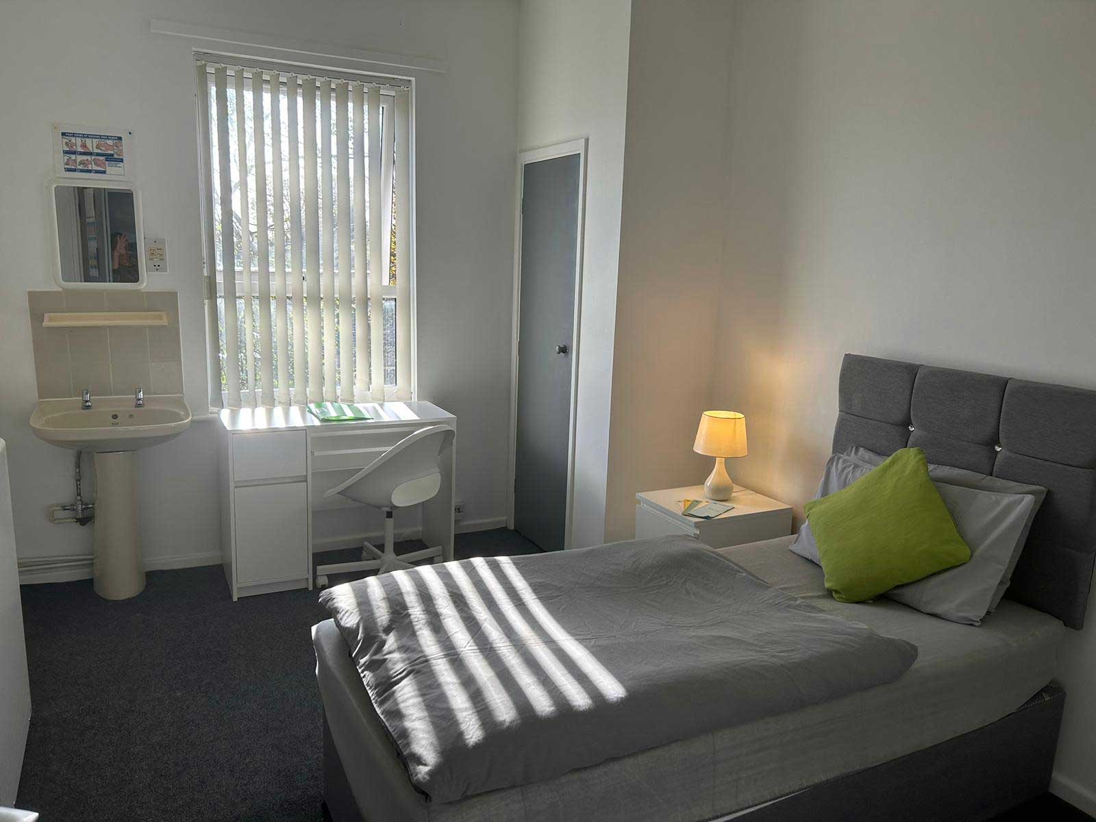 A single bed in a room with a window, bedside table and a small sink