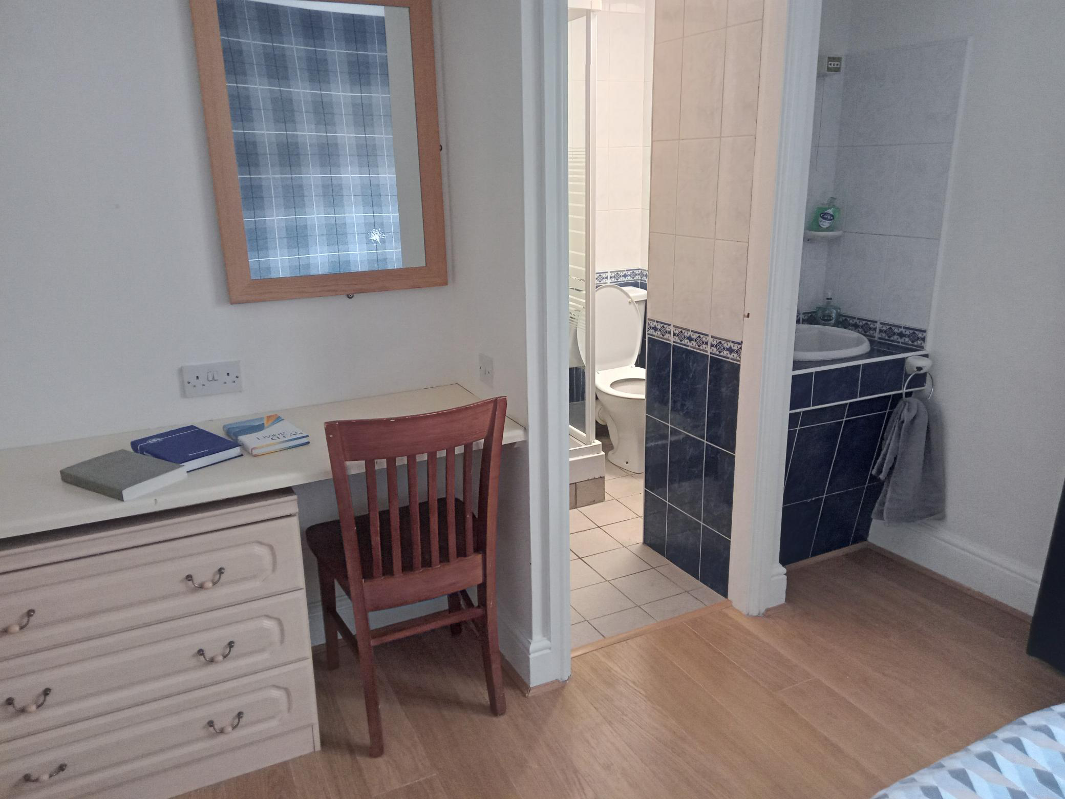 A desk  is in the corner of the room next to a door that leads to a bathroom. On the other side of the doorframe is a sink basin.
