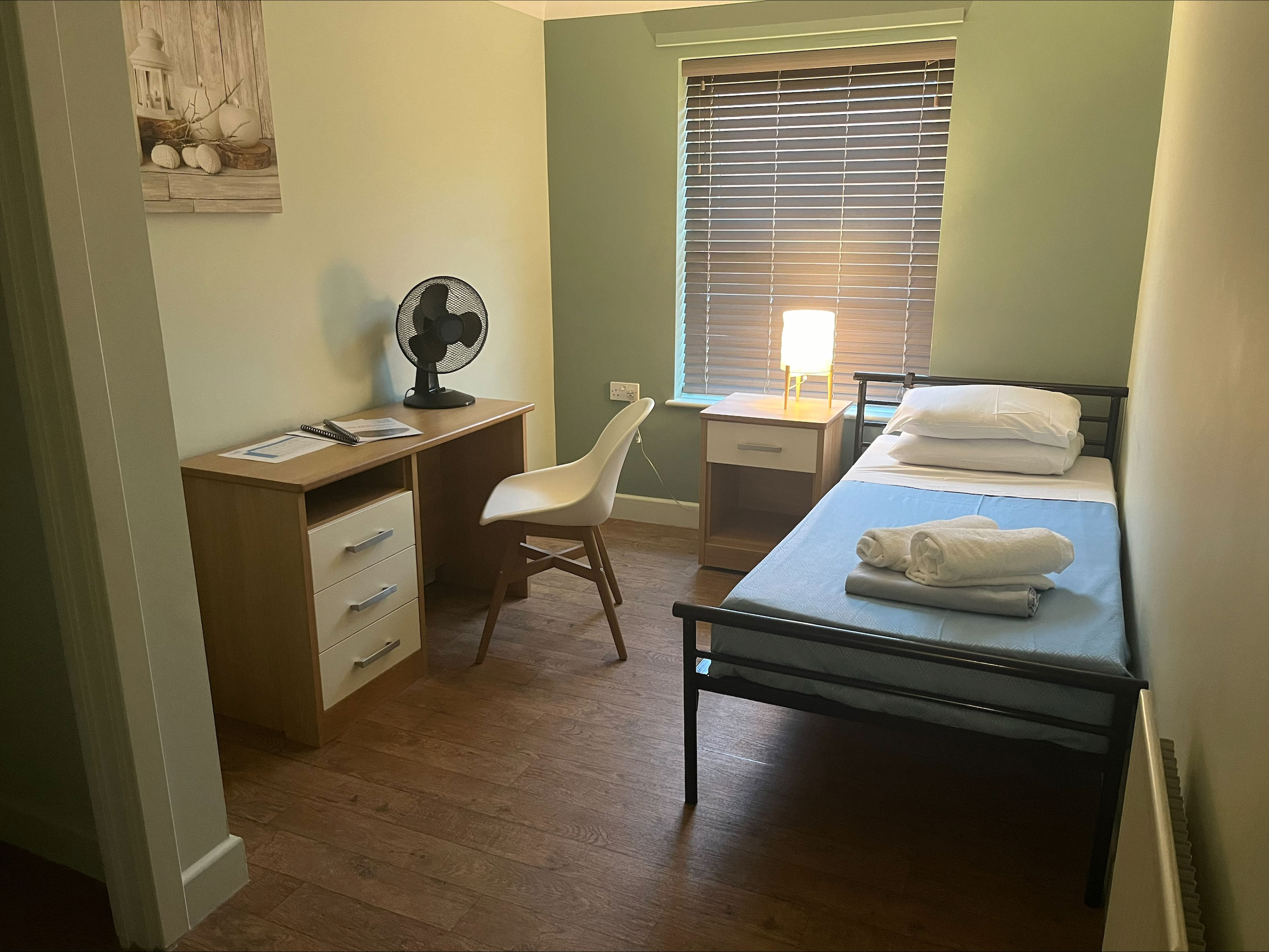 A single bed next to the wall with a desk, chair and shelf opposite