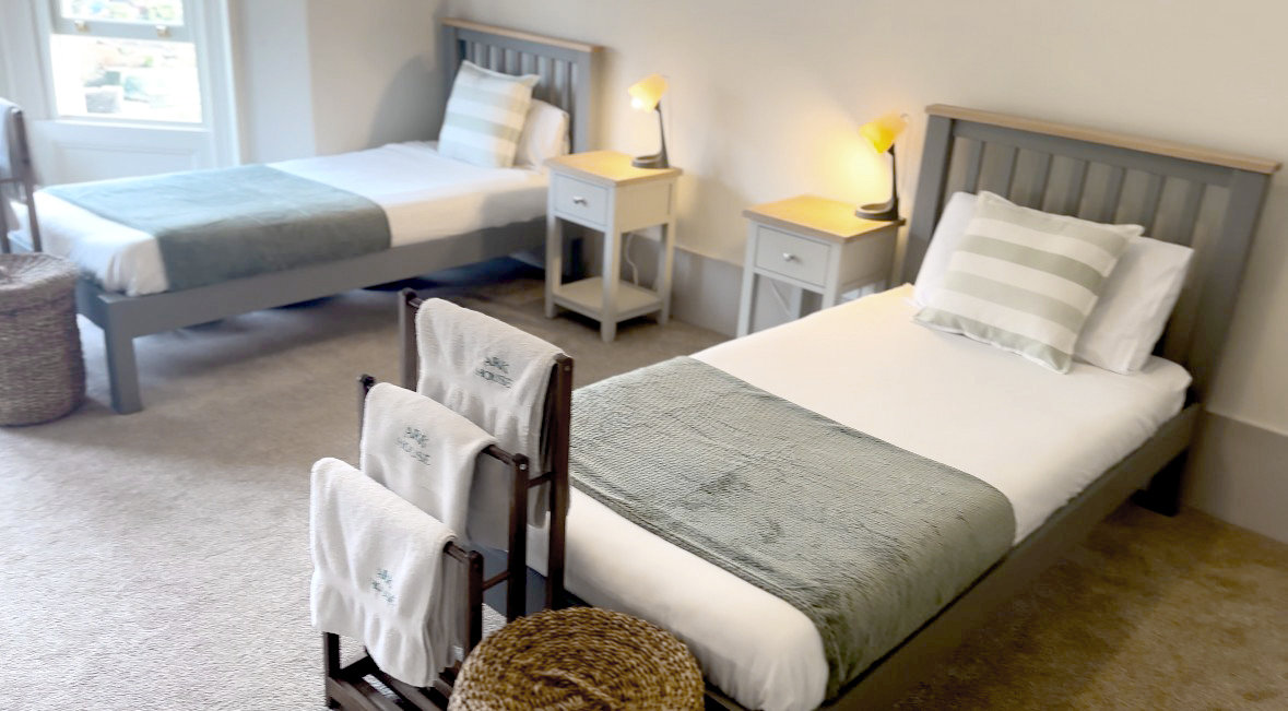 Two single beds, each with a basket and towels at the foot of the bed and a bedside table with a lamp