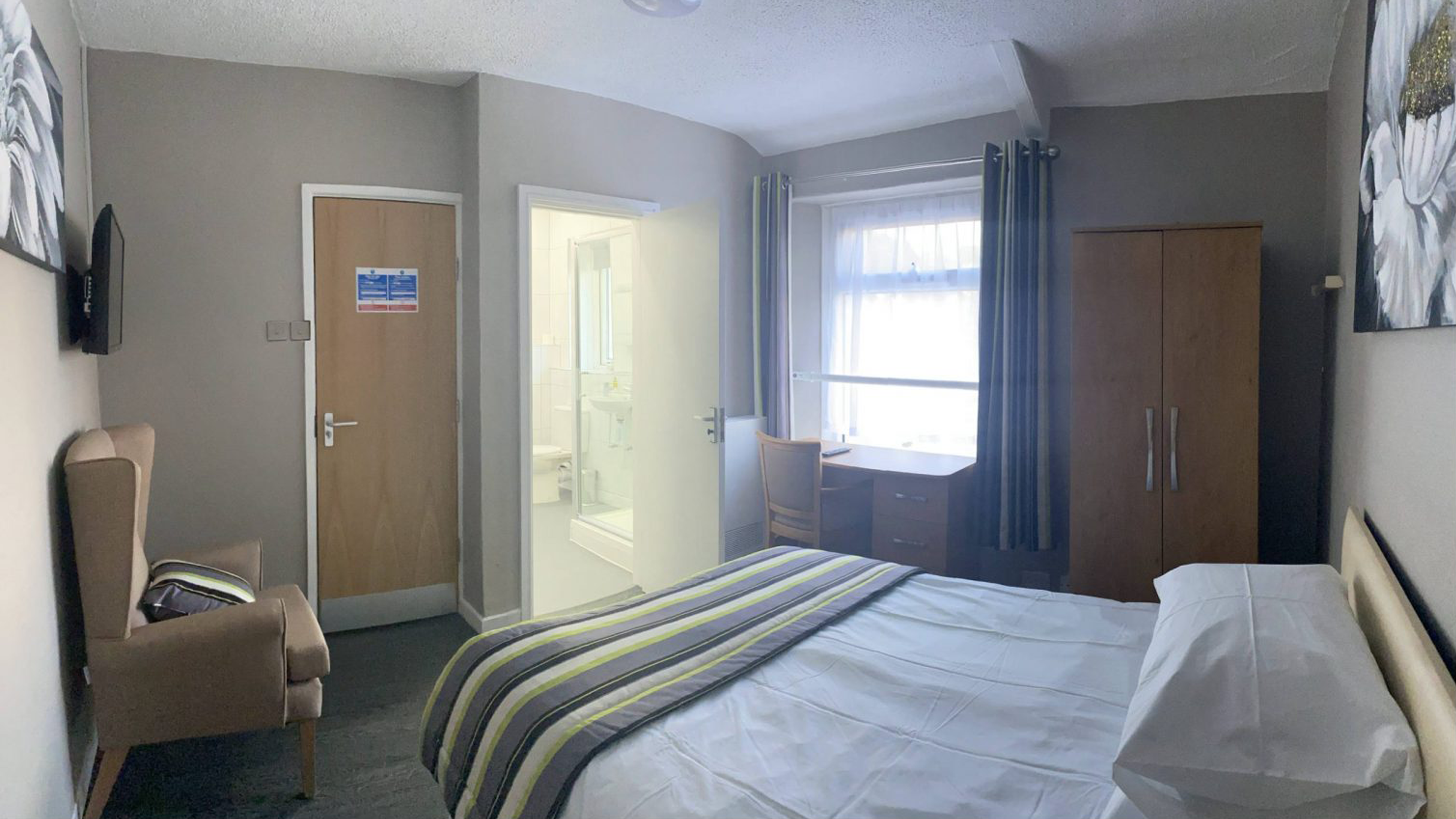 A bedroom with an en suite bathroom. The room contains an arm chair, wardrobe, desk, TV, and bed