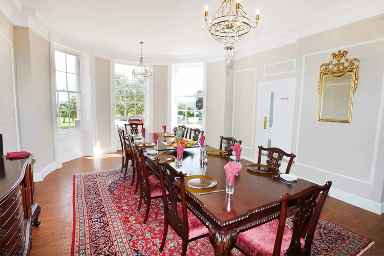 A long dark wood dining table with matching chairs tucked in. A red rug sits under the table and a bay window looks out onto the grounds