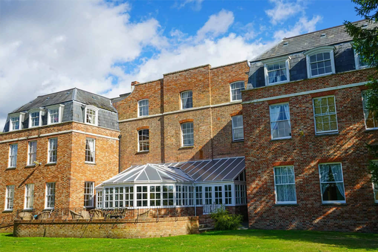 A brick exterior showing the back of a manor house. A glass conservatory is attached along with outside seating on a patio leading to the vast garden.