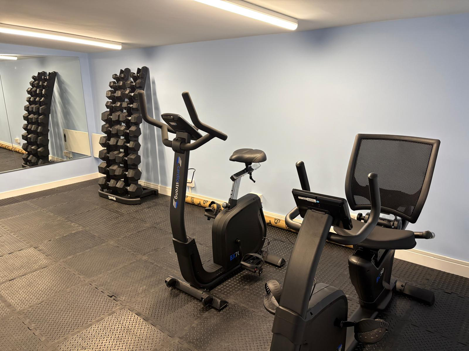 A small gym room with a stack of dumbbells, two exercise bikes and a mirrored wall