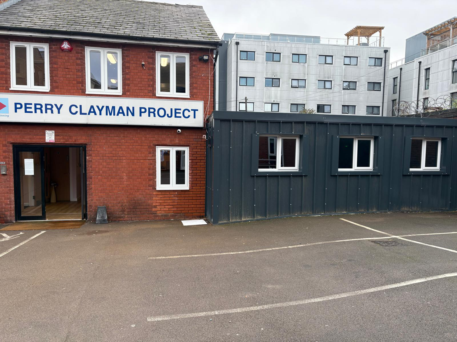 The outside of a brick building, a sign of Perry Clayman Project is above the door and there is a dark green converted shipping container next to it