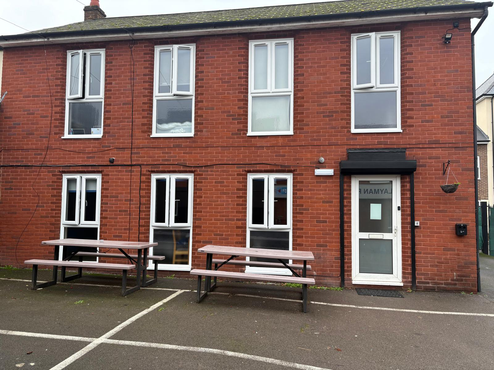 A brick exterior with lots of windows and a grey courtyard. There are 2 wooden picnic tables against the wall