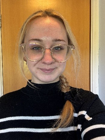 Becki is wearing glasses and a black turtleneck top. Her blonde hair is plaited and draped over her shoulder