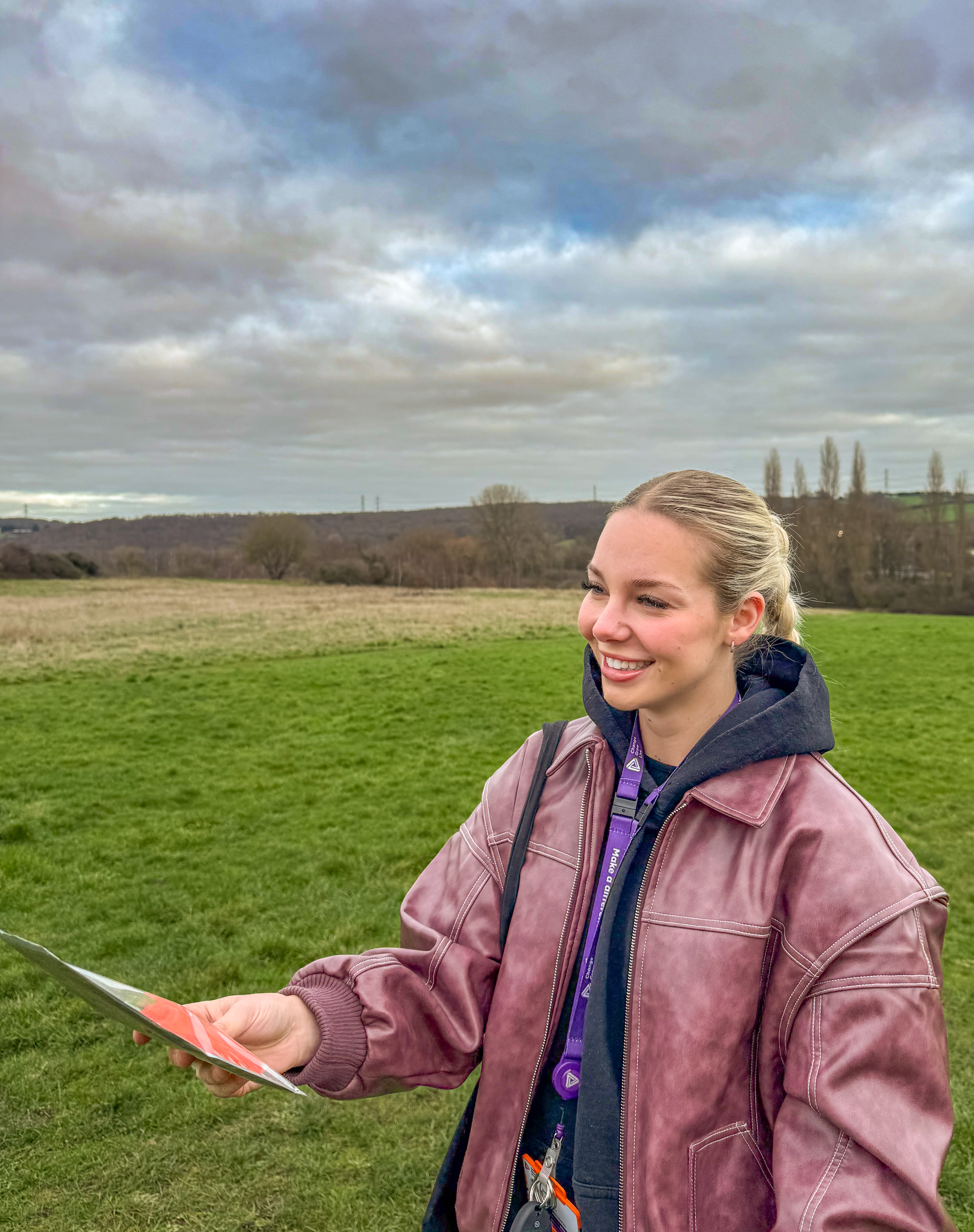 A young woman wearing a jacket and hoodie smiles while holding a leaflet in an open field, with trees and hills in the background beneath a cloudy sky.