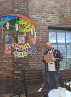 Fran is stood in front of a brick wall. He is holding brown clipboard and stood next to a rainbow umbrella