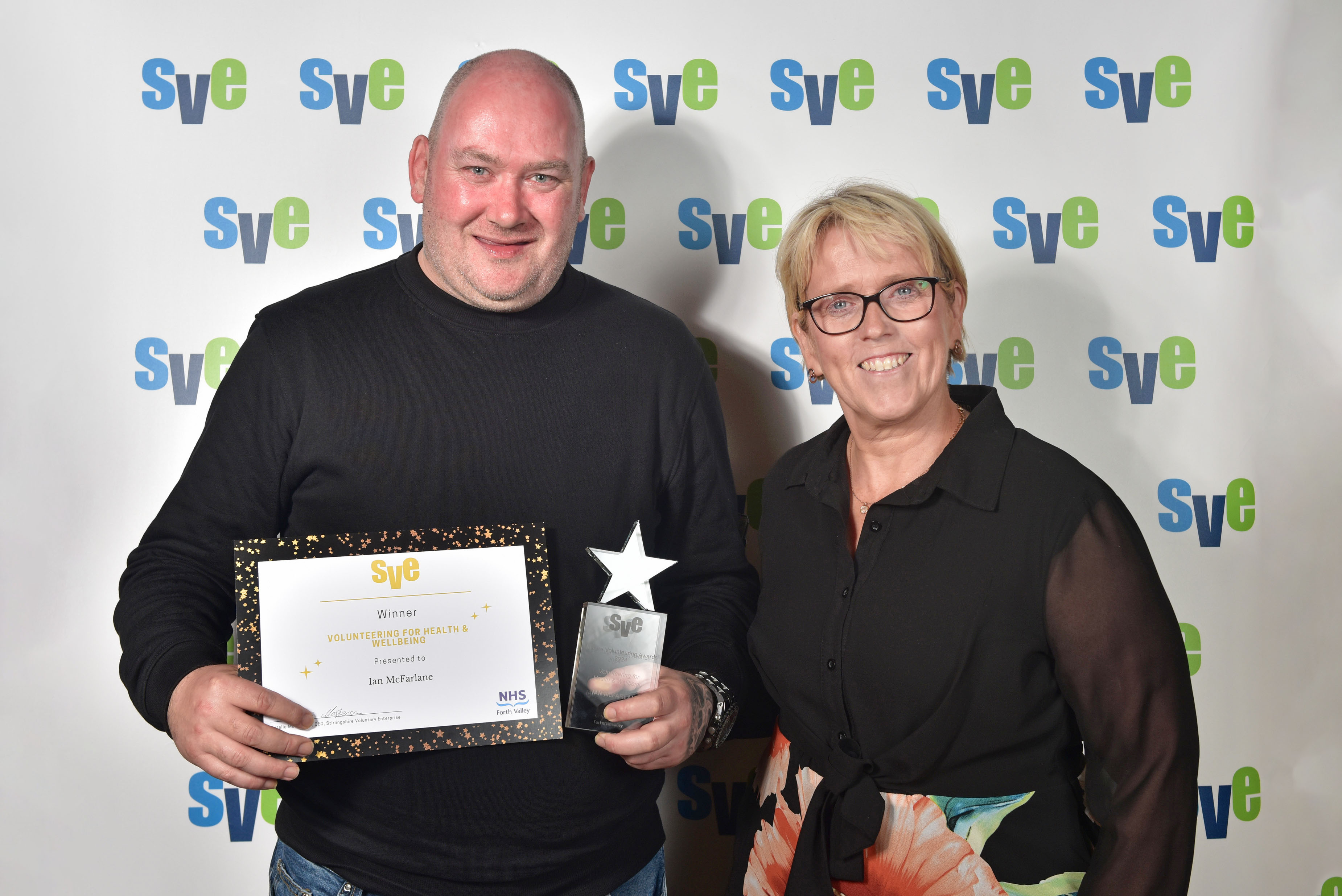 A man and a woman stood smiling. The man is holding up a certificate and an award.