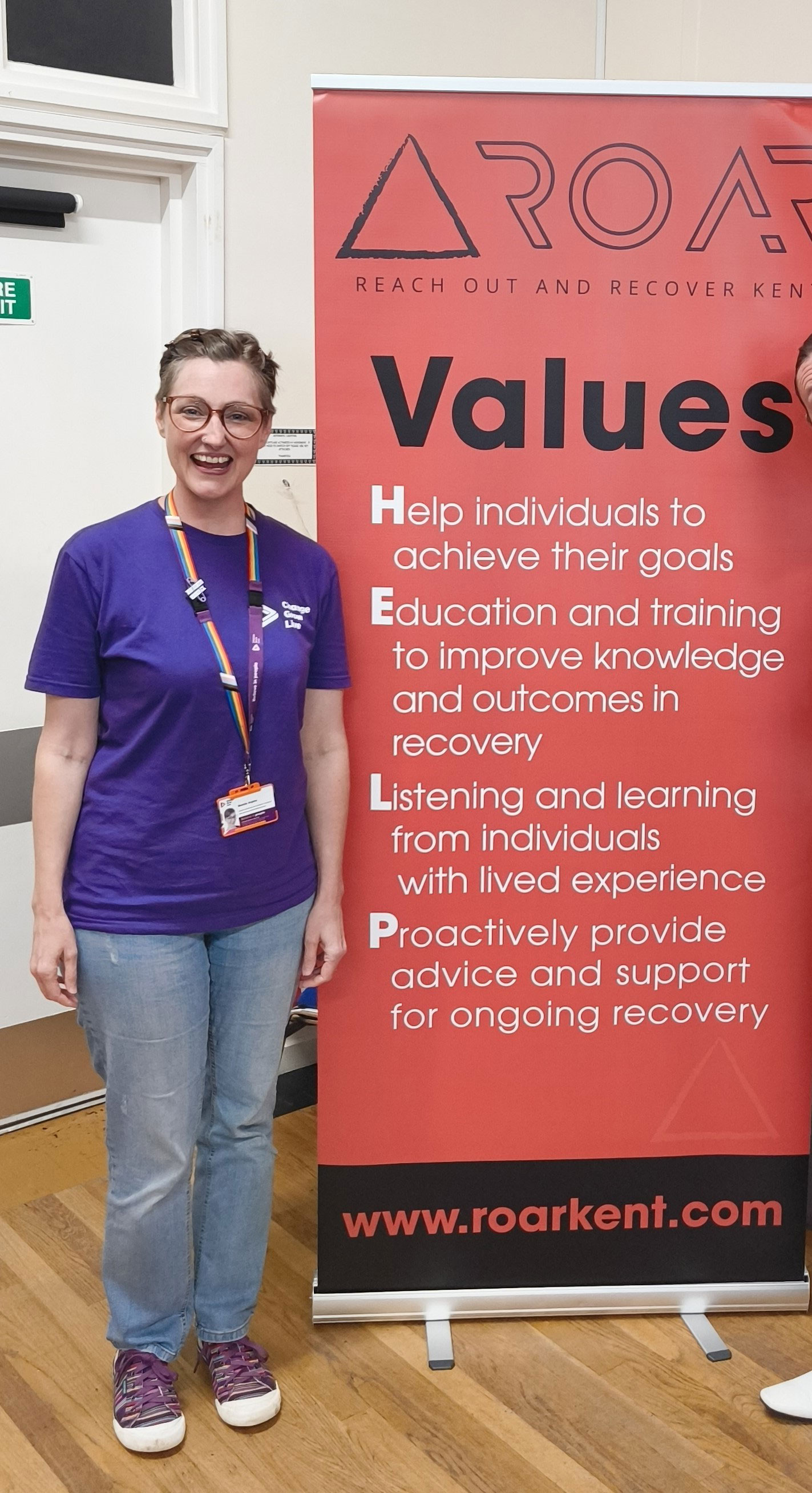 Bonnie is stood wearing a purple t-shirt and jeans, standing next to a roller banner with information about volunteering