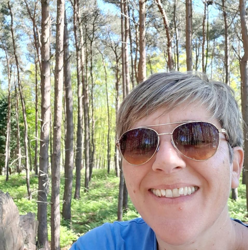 A woman with short cropped hair, wearing sunglasses and stood in a forest of trees.