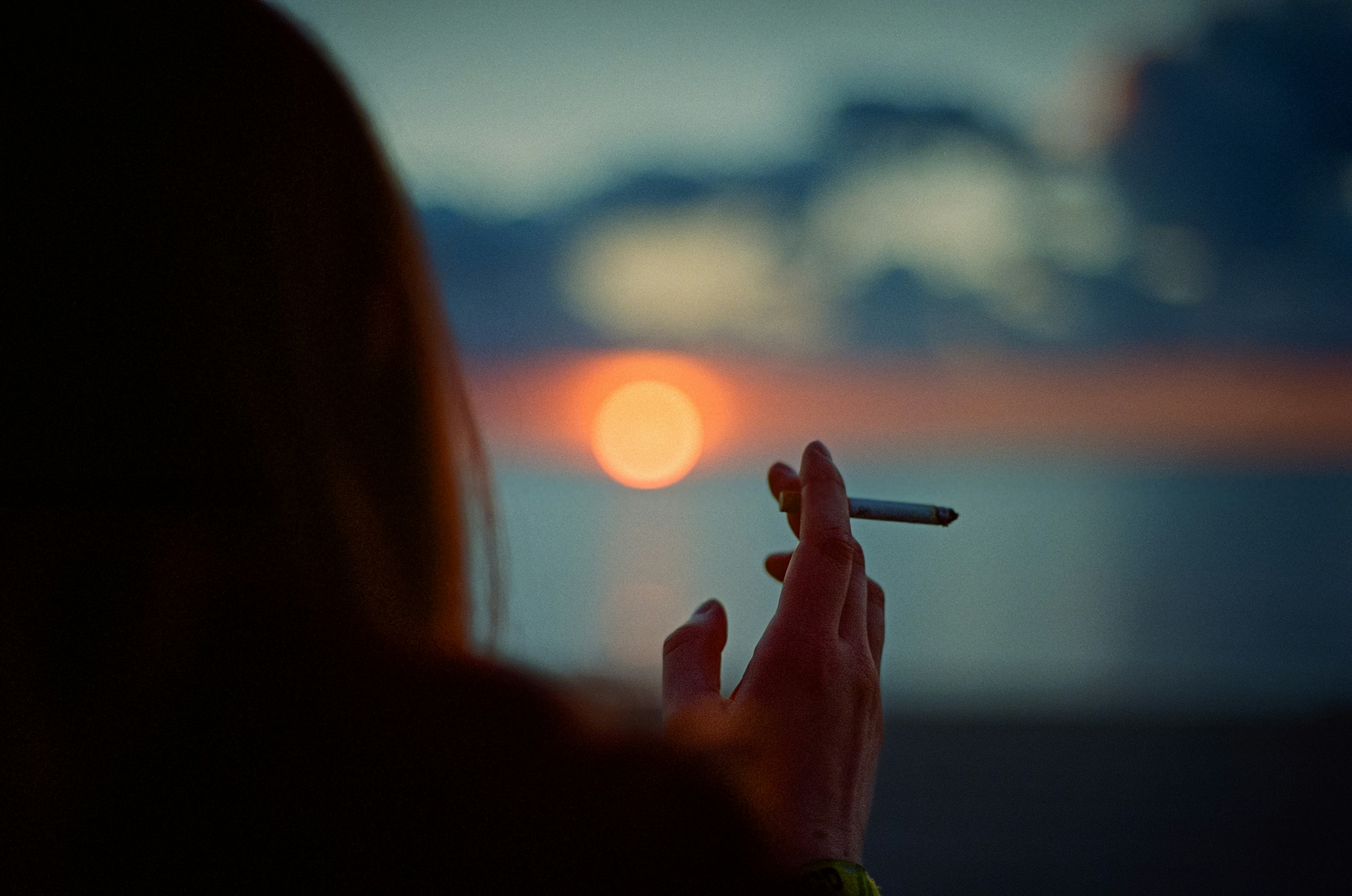 The back of a women smoking against a summer sunset over water.