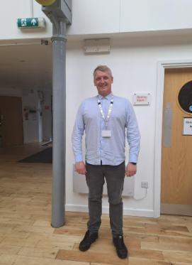 Craig is wearing a light coloured shirt and grey trousers, stood in front of a white wall.