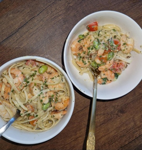 A wooden table with two white bowls of linguine prawn pasta and silver spoons coming out of the bowls.