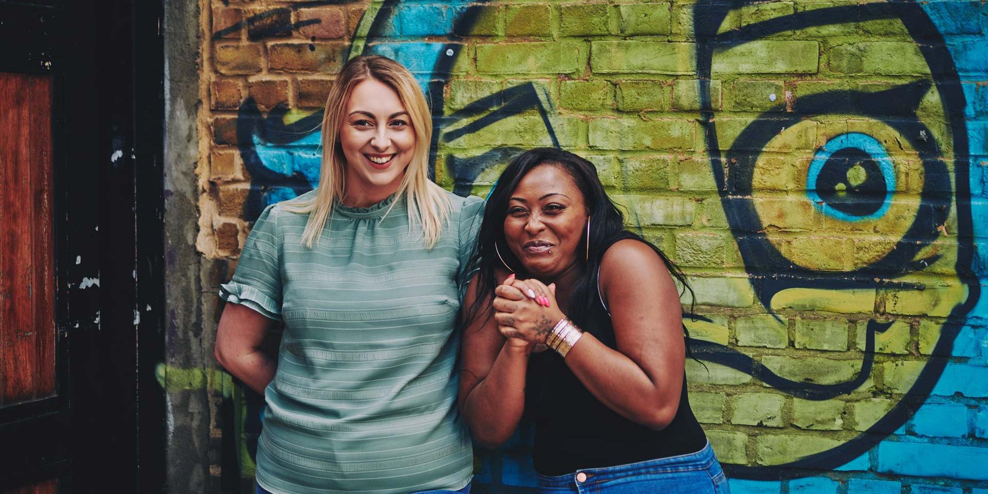 two women laughing together standing in front of some street art