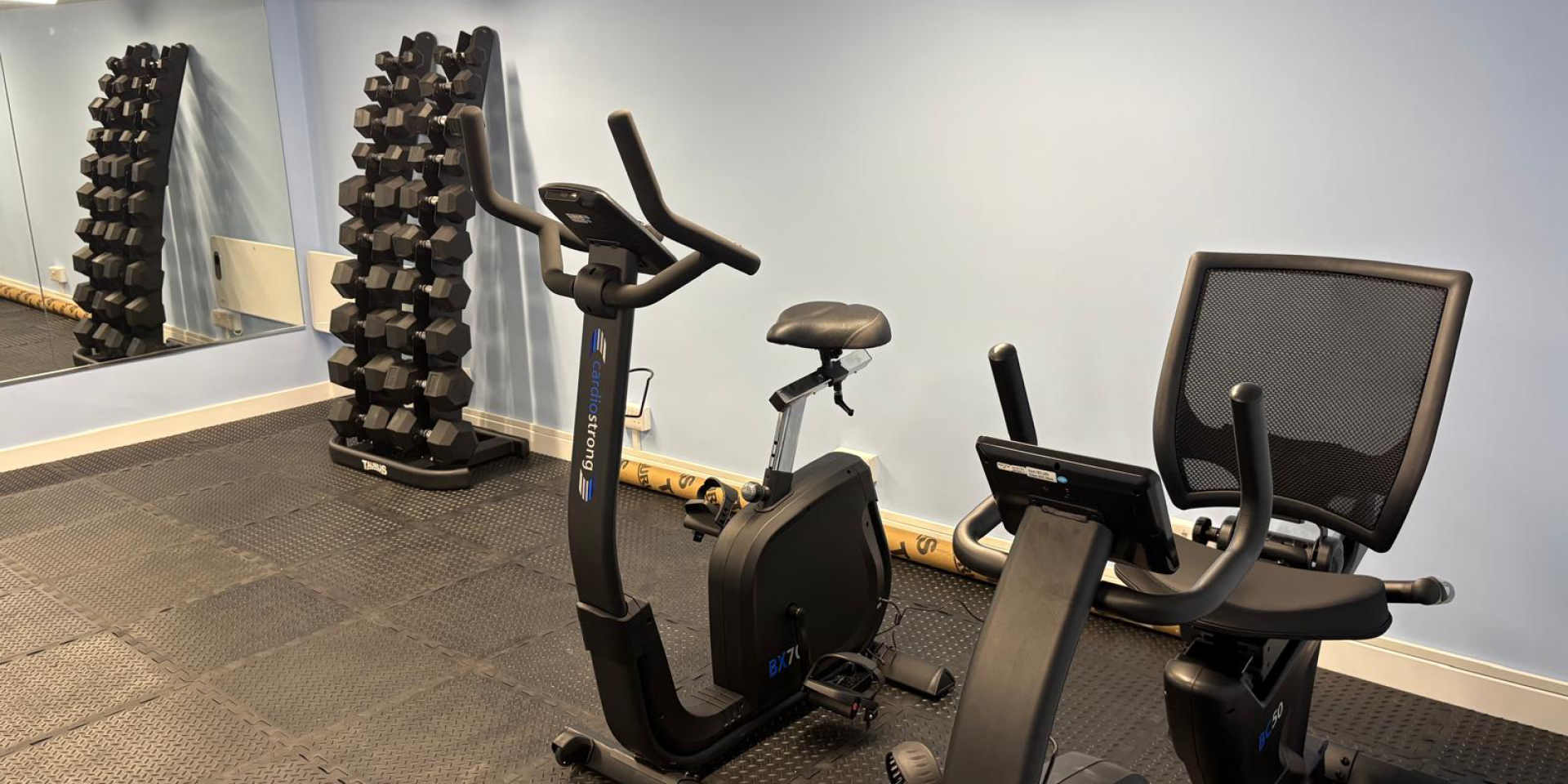 A small gym room with a stack of dumbbells, two exercise bikes and a mirrored wall