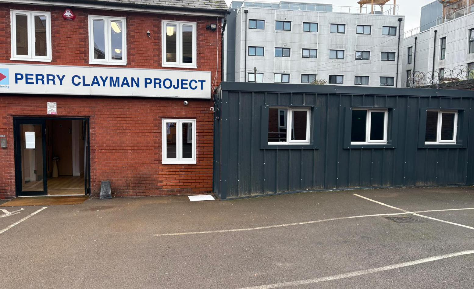 The outside of a brick building, a sign of Perry Clayman Project is above the door and there is a dark green converted shipping container next to it