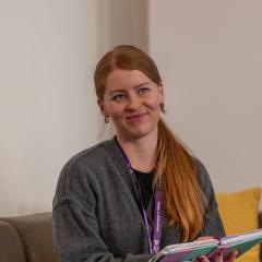 A young woman with ginger hair in a ponytail over her shoulder is sat on a sofa looking at someone off camera