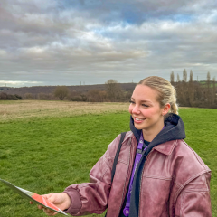 A young woman wearing a jacket and hoodie smiles while holding a leaflet in an open field, with trees and hills in the background beneath a cloudy sky.