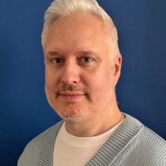 Lee has white hair and is wearing a blue v neck jumper over a white t-shirt
