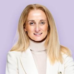 Victoria has long blonde hair and is wearing a camel coloured turtleneck top and white jacket