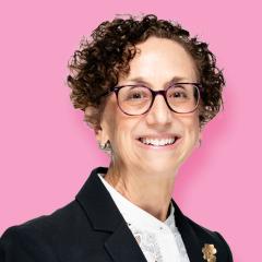 Deborah has short curly hair and is wearing glasses, a white shirt and black jacket