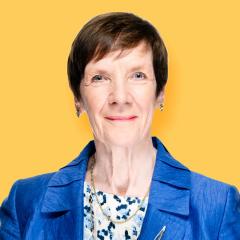 Dr Caroline has a pixie cut brown hair, and is wearing a floral top with blue jacket