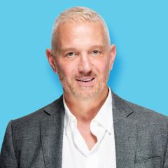 Michael has short blonde hair, beard and mustache. He is wearing a white shirt with the top buttons open and a grey blazer