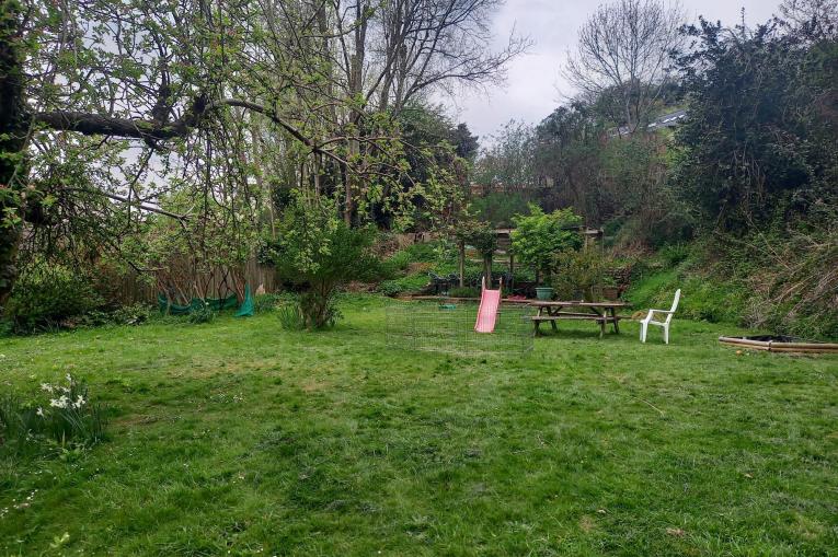 A photo of a big garden. There is plenty of open space and greenery, including grass and trees. There is a child's slide and a wooden picnic bench towards the back of the garden. 