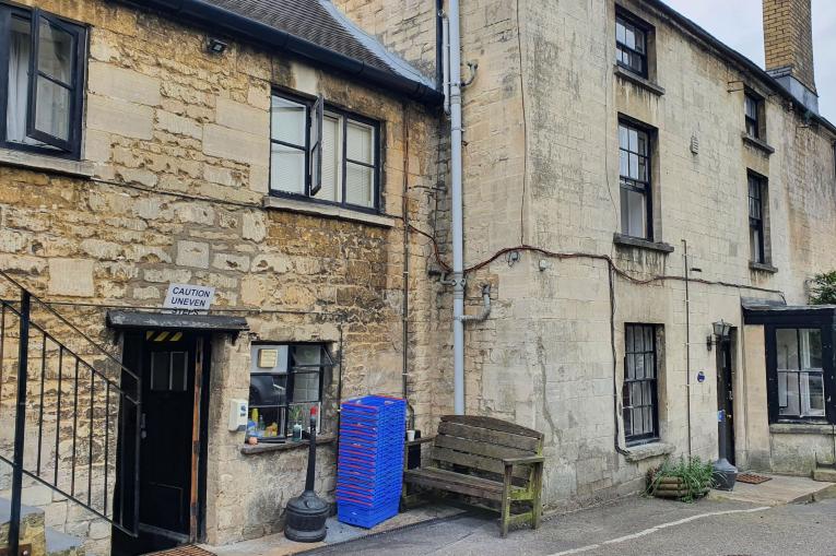 A photo of an older building made of stone. There are steps leading up to another level to the left of the photo and a door at the bottom with a sign above it that reads 'Caution uneven steps'. There is a wooden bench against the wall of the building. 