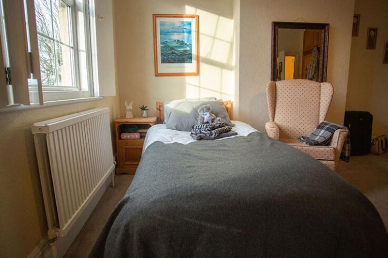 A photo of a bedroom. There is a single bed in the middle of the image. On the wall to the left of the bed is a radiator and a bedside table. Above the radiator is a window with shutters. An arm chair sits to the right of the bed in front of a big mirror. 