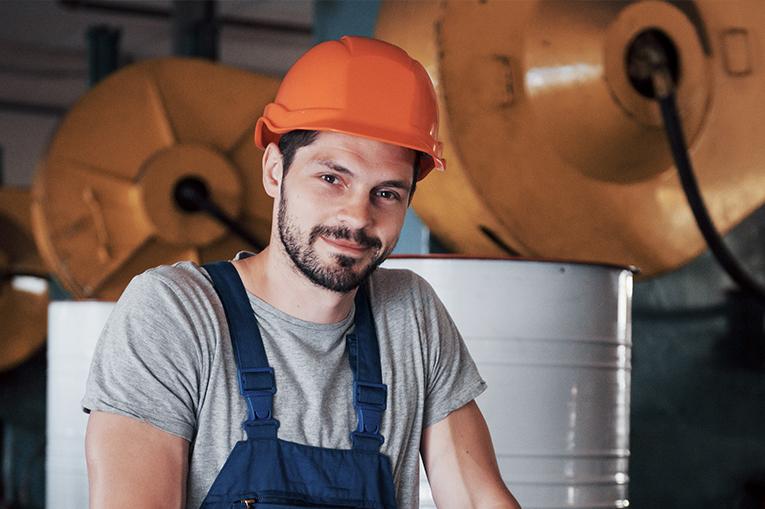 A man wearing an orange hard had and blue overalls stood in front of machinery